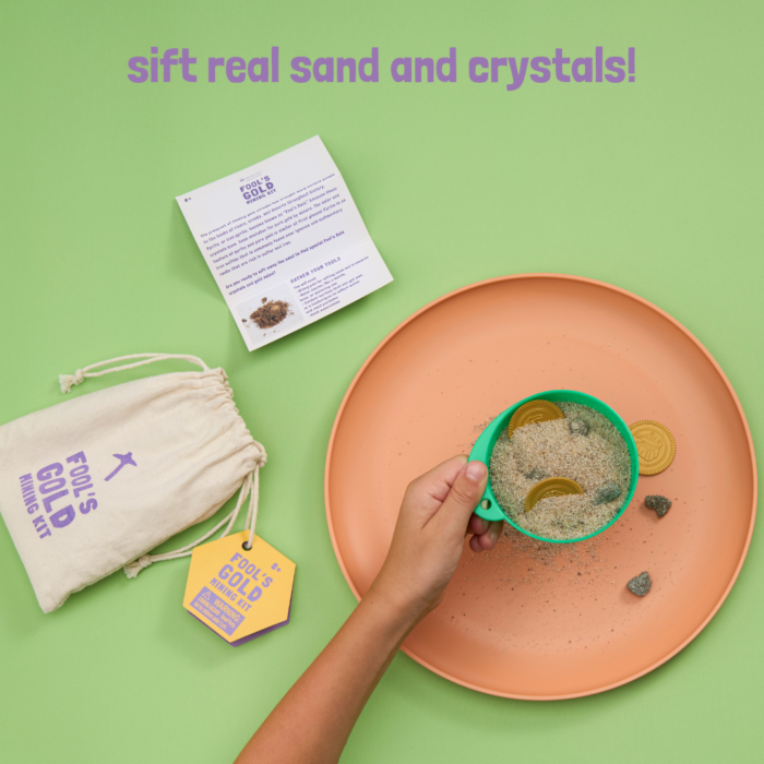 A child's hand uses a green sifter to sift sand, plastic gold coins, and pyrite crystals. A muslin bag with the text wholesale fool's gold mining kit and an instruction pamphlet is present