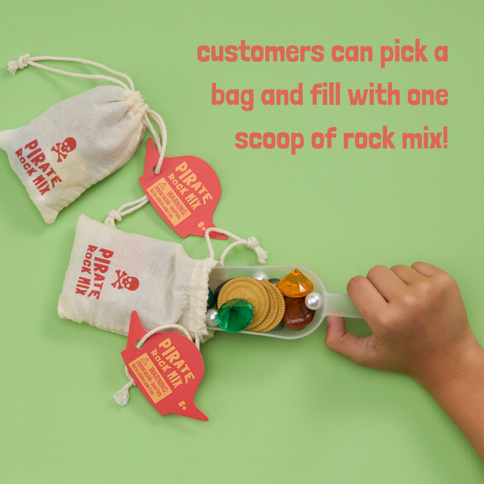 A child's hand scooping acrylic gems, plastic gold coins, plastic pearls, and tumbled crystals into a bag with the text "Pirate Rock Mix." Text: "customers can pick a bag and fill it with one scoop of wholesale rock mix!"
