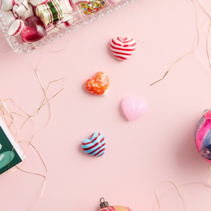 wholesale artisan glass hearts on a pink background. hearts are multicolored with various patterns. there is holiday tinsel, candies, and ornaments peeking in from the sides of the image