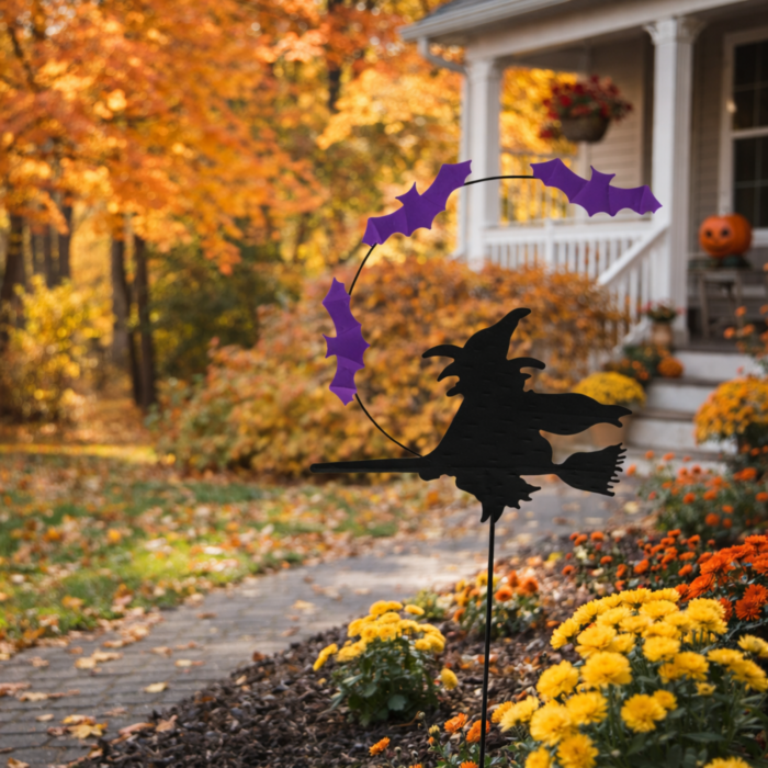 wholesale halloween garden stake showing the black silhouette of a witch flying on a broom with purple bats flying overhead. this is shown in a front yard garden during the fall with a white porch and orange leaves behind it