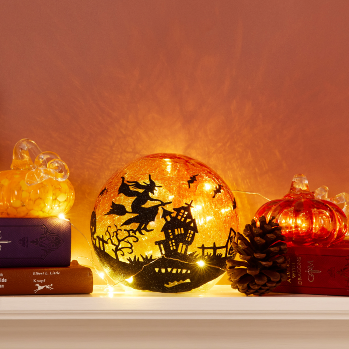 wholesale crackle glass LED lantern showing the black silhouette of a witch on a broomstick flying over a landscape featuring a whimsical crooked haunted house, crooked trees, and bats flying around her. The background is dark orange crackle glass and shows a light orange full moon behind the witch. This is shown on a mantle decorated for halloween surrounded by pine cones and glass pumpkins.