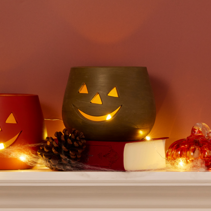 orange clay jack o lantern candle holder lit from within shown on a mantle decorated for halloween with spider webs, pine cones, glass pumpkins, and other jack o lanterns.
