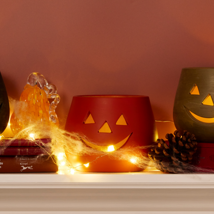 orange clay jack o lantern candle holder lit from within shown on a mantle decorated for halloween with spider webs, pine cones, glass pumpkins, and other jack o lanterns.