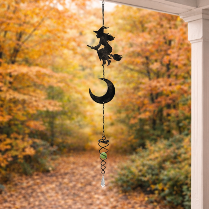 wholesale glow in the dark hanging garden wind spinner featuring a witch on a broomstick and a crescent moon. this is shown hanging on a porch facing out onto an autumnal forest